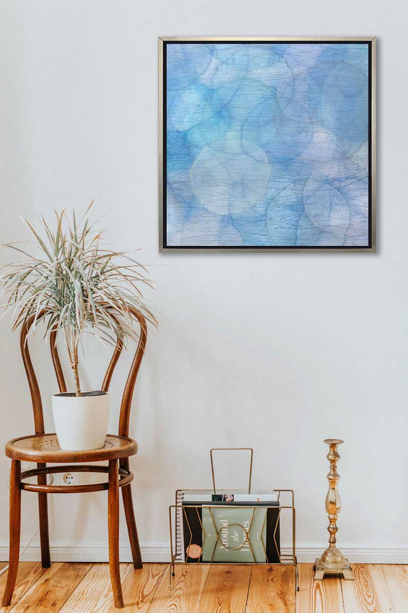 Layers of overlapping circles with an iridescent feel in blues, light purples and light greens staged in a silver floater frame above a chair and magazine rack.