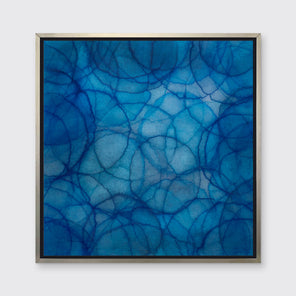 A dark and light blue abstract overlapping circle print in a silver floater frame hangs on a white wall.
