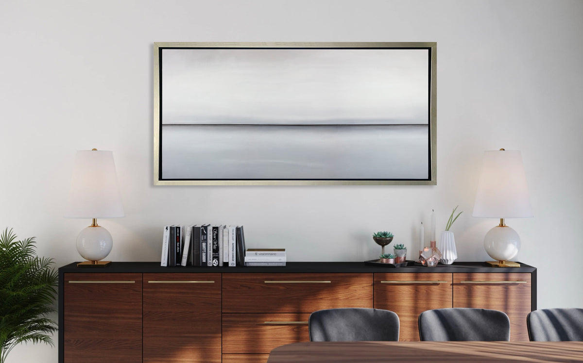 A light grey toned abstract landscape print in a silver floater frame hangs on a white wall above a dark wood credenza with a stack of books, some decorative floral accessories and two matching white table lamps.