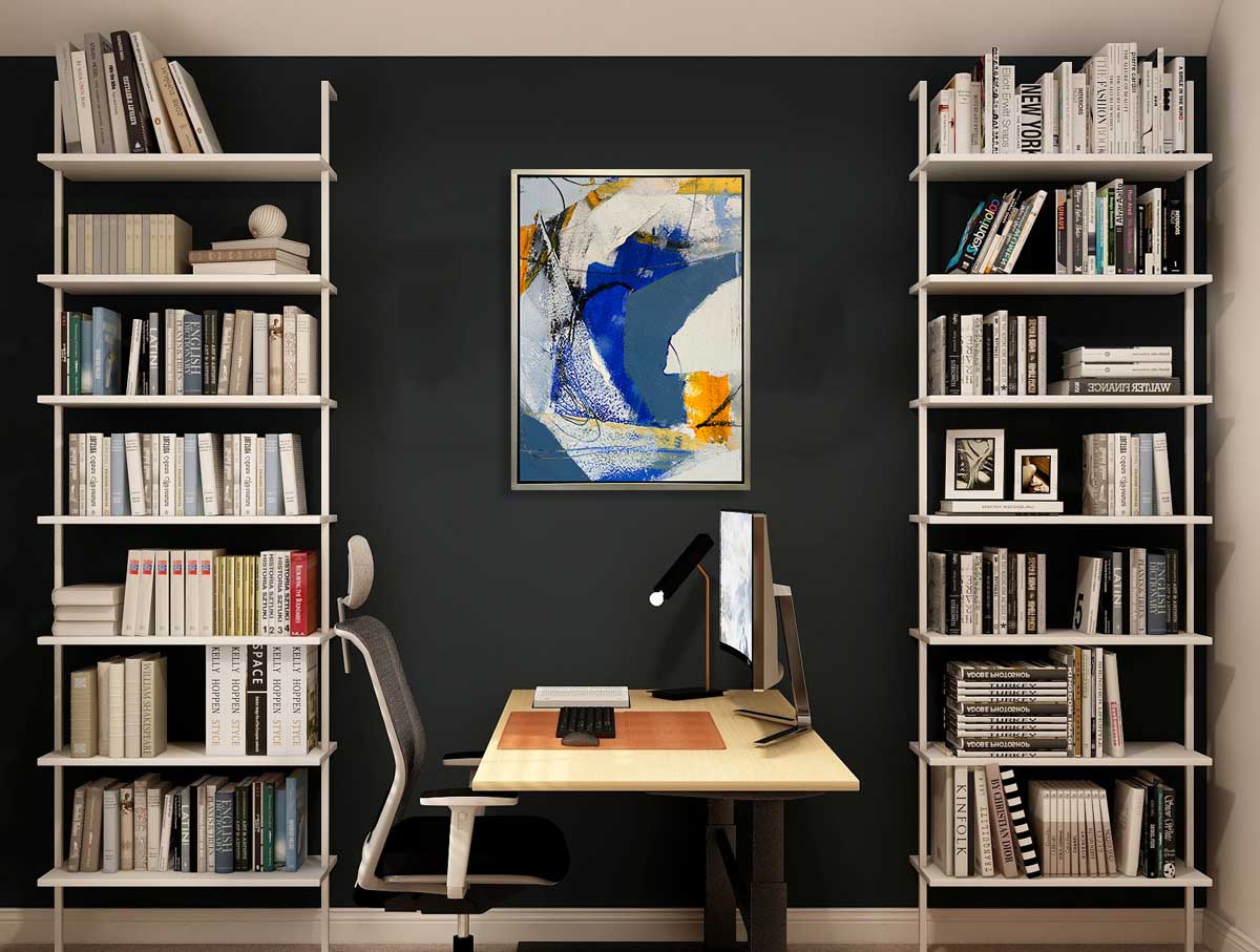 A blue, grey and yellow abstract print in a silver floater frame hangs on a charcoal wall in between two ceiling to floor bookshelves and above a desk with a computer and desk lamp.