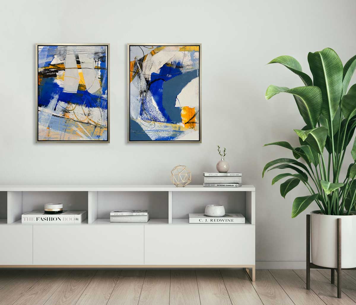 Two blue, grey and yellow abstract prints in silver floater frames hang side by side on a white wall above a white console with books, a gold wire sphere and a small vase with greenery stems.