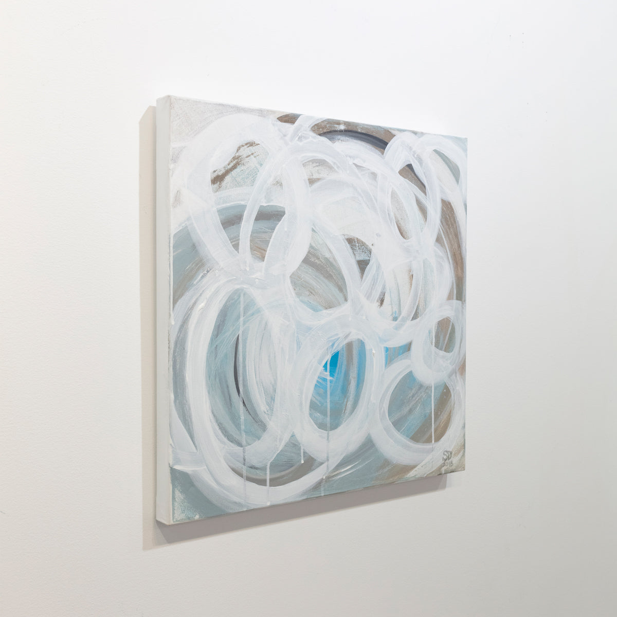 A grey, tan and blue painting with white concentric circles painted on it is seen at an angle on a gallery wall. Wired and ready to hang.