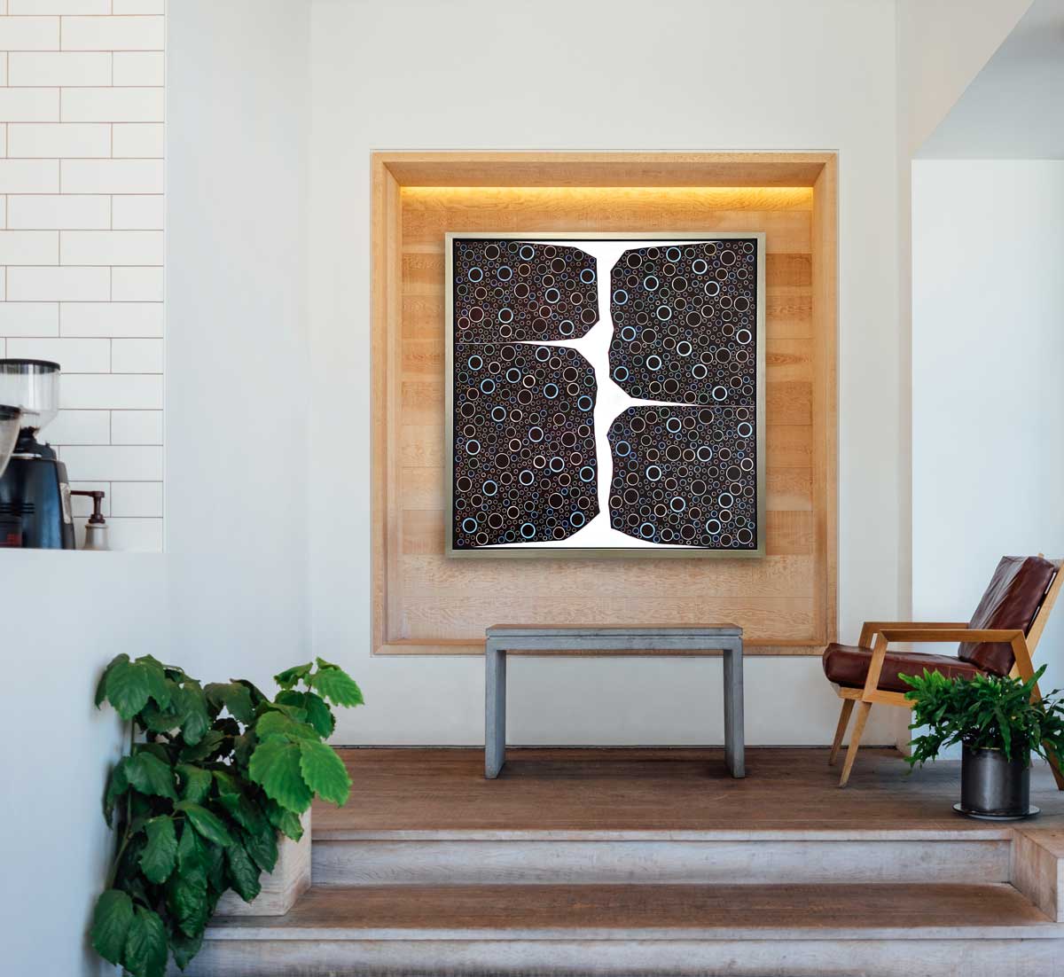 A multicolored abstract print in a silver floater frame hangs on a wood cutout above a bench and next to a leather chair..