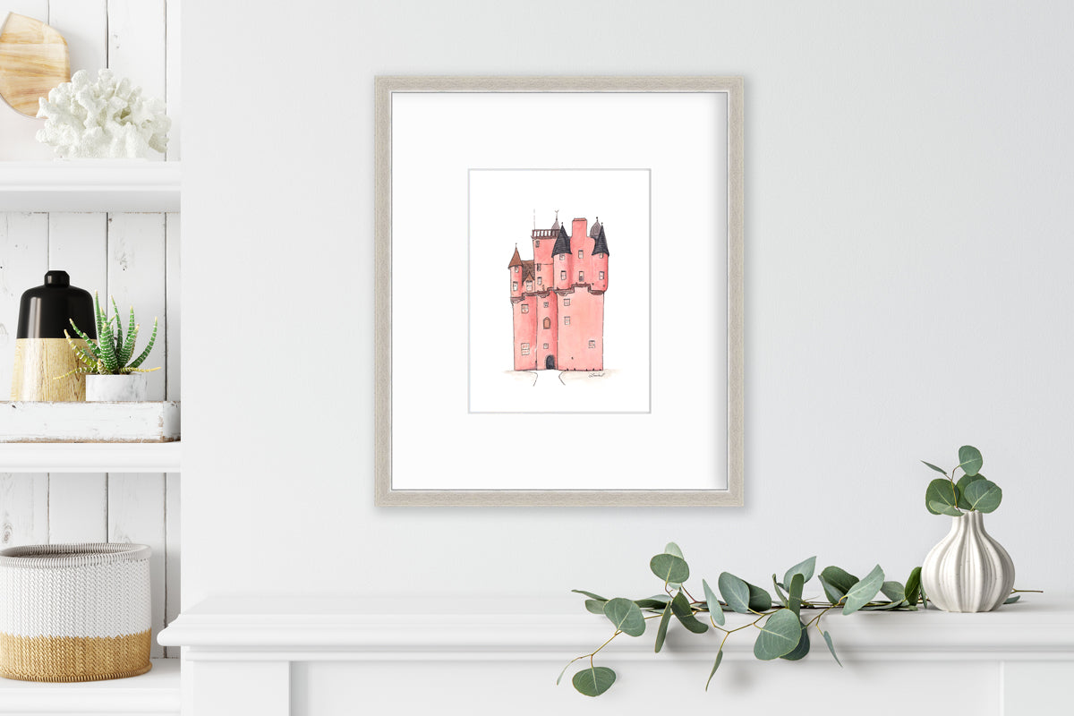 A pink and grey castle print in a silver frame with a mat hangs on a white wall above a white shelf with a eucalyptus filled white vase and next to a built in bookshelf with decorative accessories.