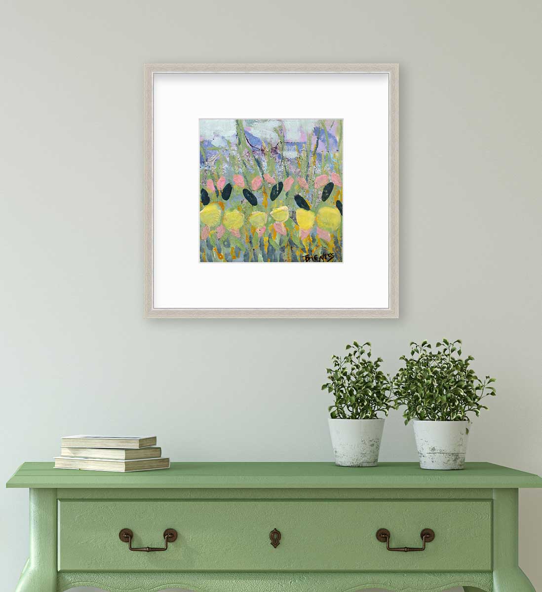 A green, yellow and pink abstract floral print in a silver frame with a mat hangs on a white wall above a green console table with a stack of books and two potted plants.