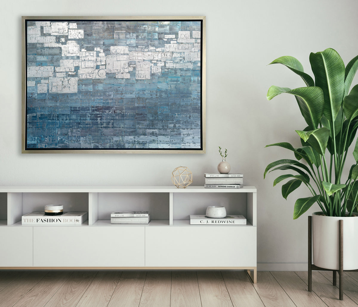 A blue, white and grey abstract print in a silver floater frame hangs on a white wall above a white credenza with stacks of books, a gold sphere decoration and a large floor potted plant.