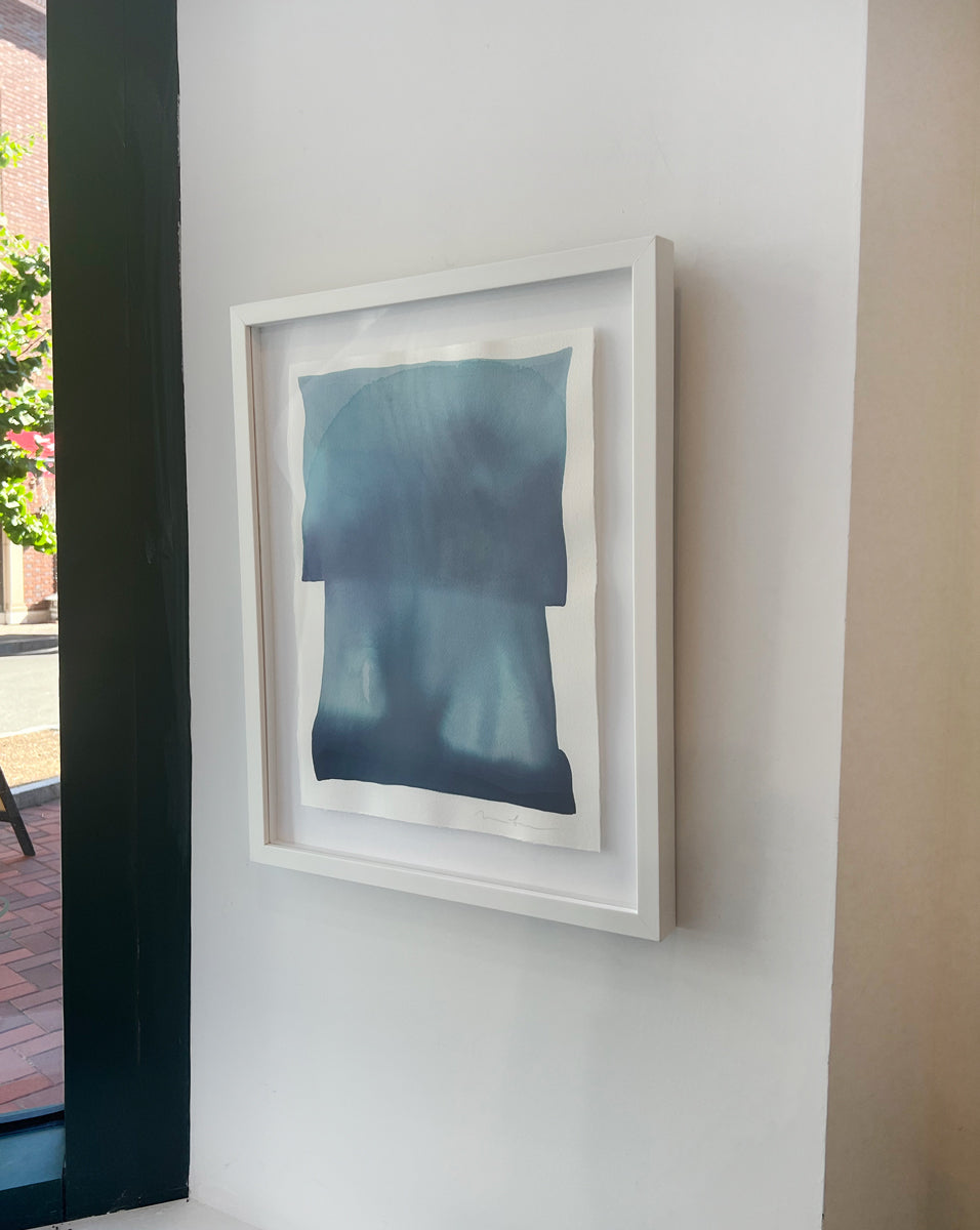 A right side-angle view of a tonal blue painting with abstract shapes by Nealy Hauschildt in a white frame floating on a white matte on a white wall.