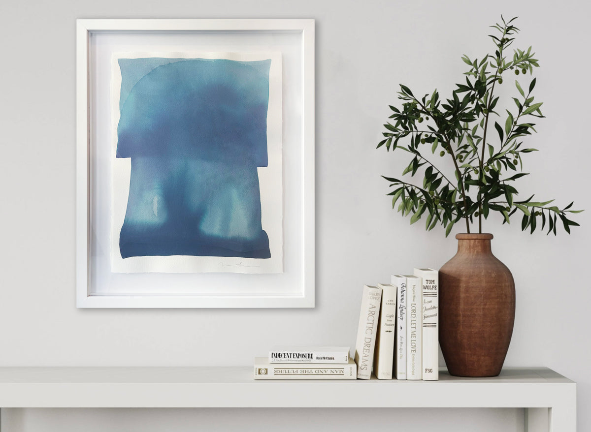 A tonal blue painting with abstract shapes by Nealy Hauschildt in a white frame floating on a white matte on a white wall above a white console table with a stack of books and a greenery filled terracotta vase.