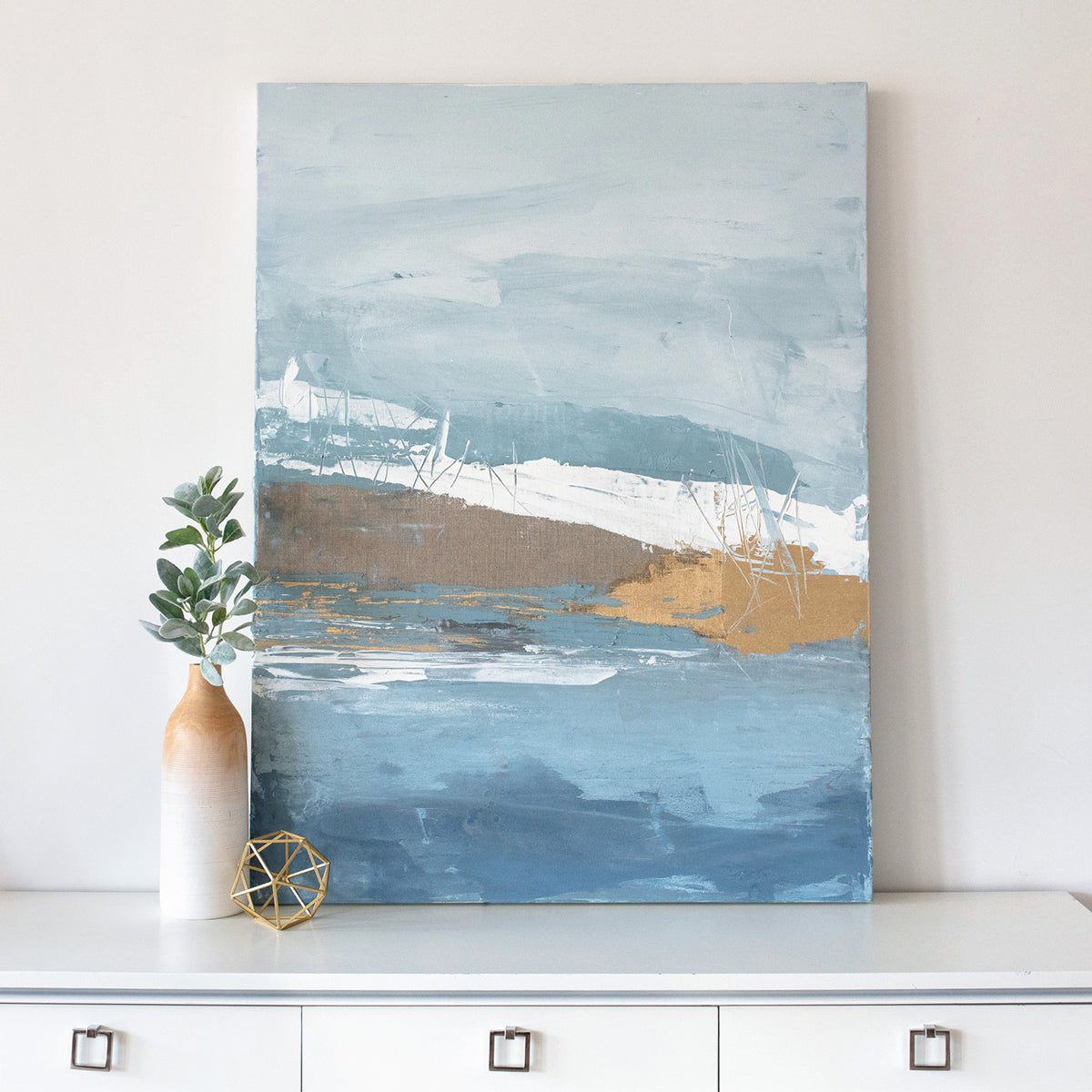 A blue, white, and gold abstract painting rests on a white dresser next to modern table decor.