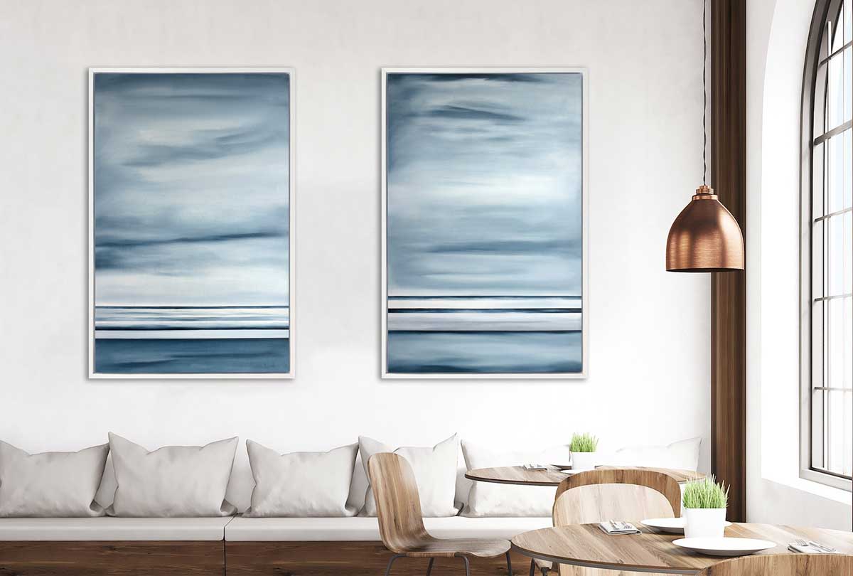 Two blue-grey abstract prints in a white floater frame on a white wall hang above a built-in bench with light grey cushions and pillows and natural wood dining tables and chairs.