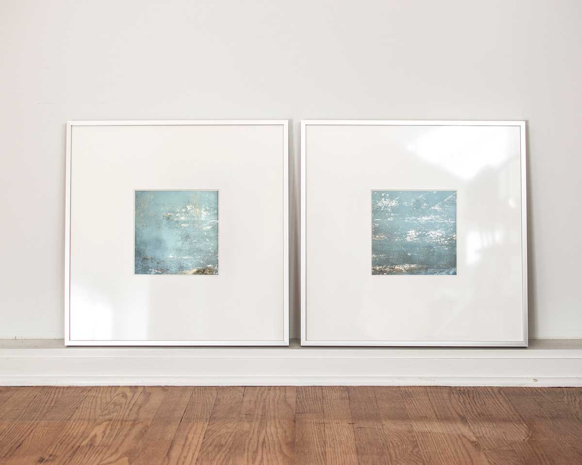 Two small light blue and silver painted art mirrors in silver frames with large white mats sit on a white surface.