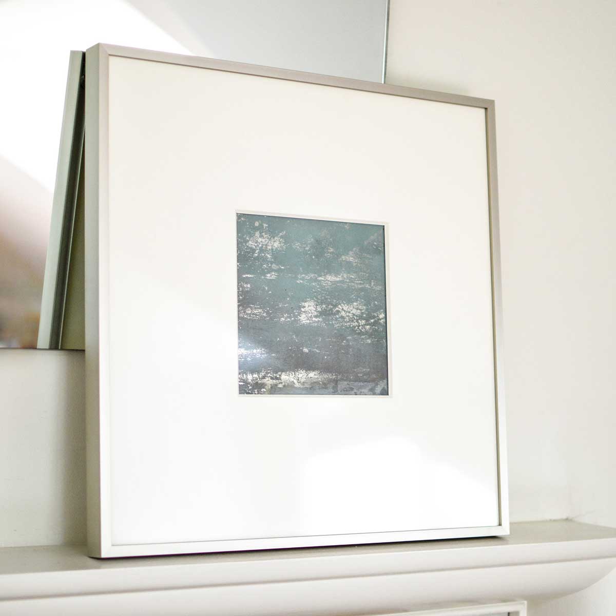 A left side-angle view of a small light blue and silver painted art mirror in a silver frame with a large white mat sits on a white surface.