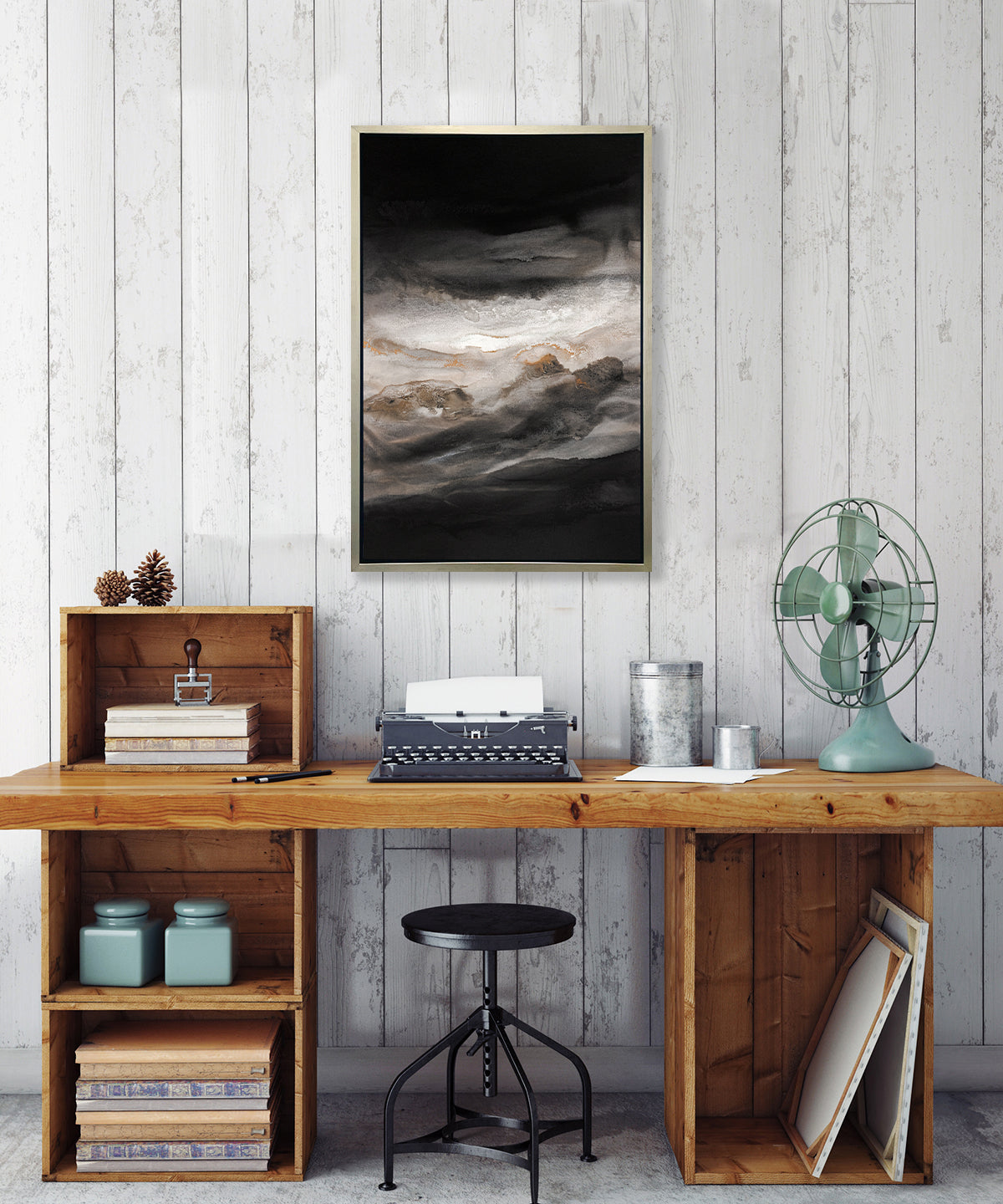 A black, white, beige and gold abstract print in a silver floater frame hangs on a white wall above a wood desk with a typewriter, two hammered silver containers, a teal table fan and a black adjustable stool.