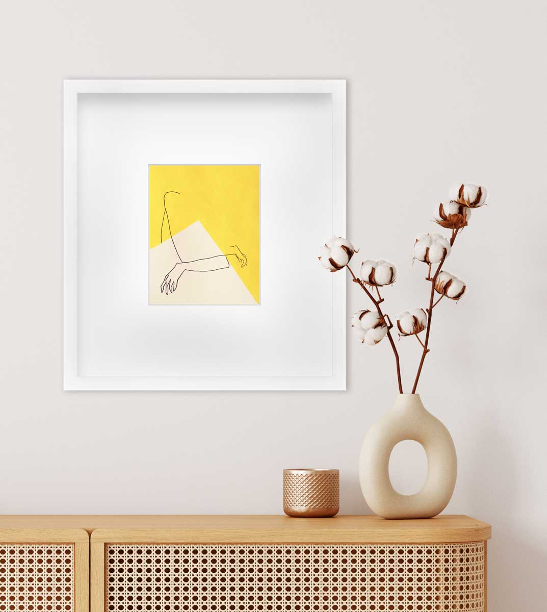A yellow and white abstract print by Hazal Ozturk in a white frame with a mat hangs on a white wall above a natural wood wicker console table with a beige and copper vases with flowers in them.