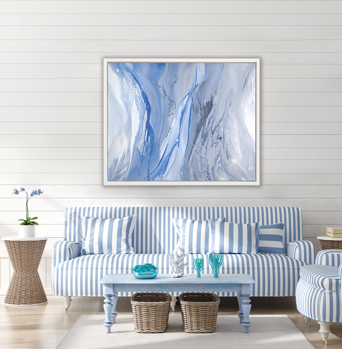 A blue, white and grey abstract print in a white floater frame hangs on a white wall above a blue and white striped sofa, a matching chair and a light blue rectangular coffee table.