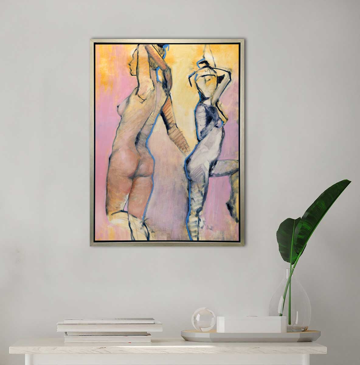 A pink, yellow, blue and black abstract figurative print of two women in a silver floater frame hangs on a white wall above a white table with a stack of books and tray with with a white box, a clear sphere paperweight and a clear vase with two large leaf stems.