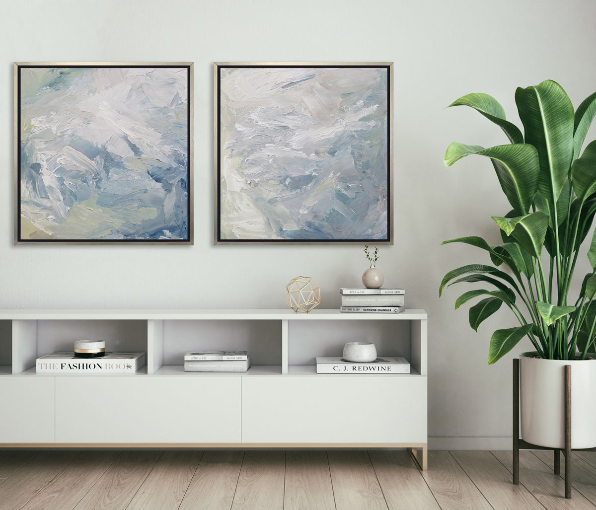 Two white, blue, grey and light green abstract prints in silver floater frames hang on a white wall above a white credenza with decorative accessories and a large potted floor plant.