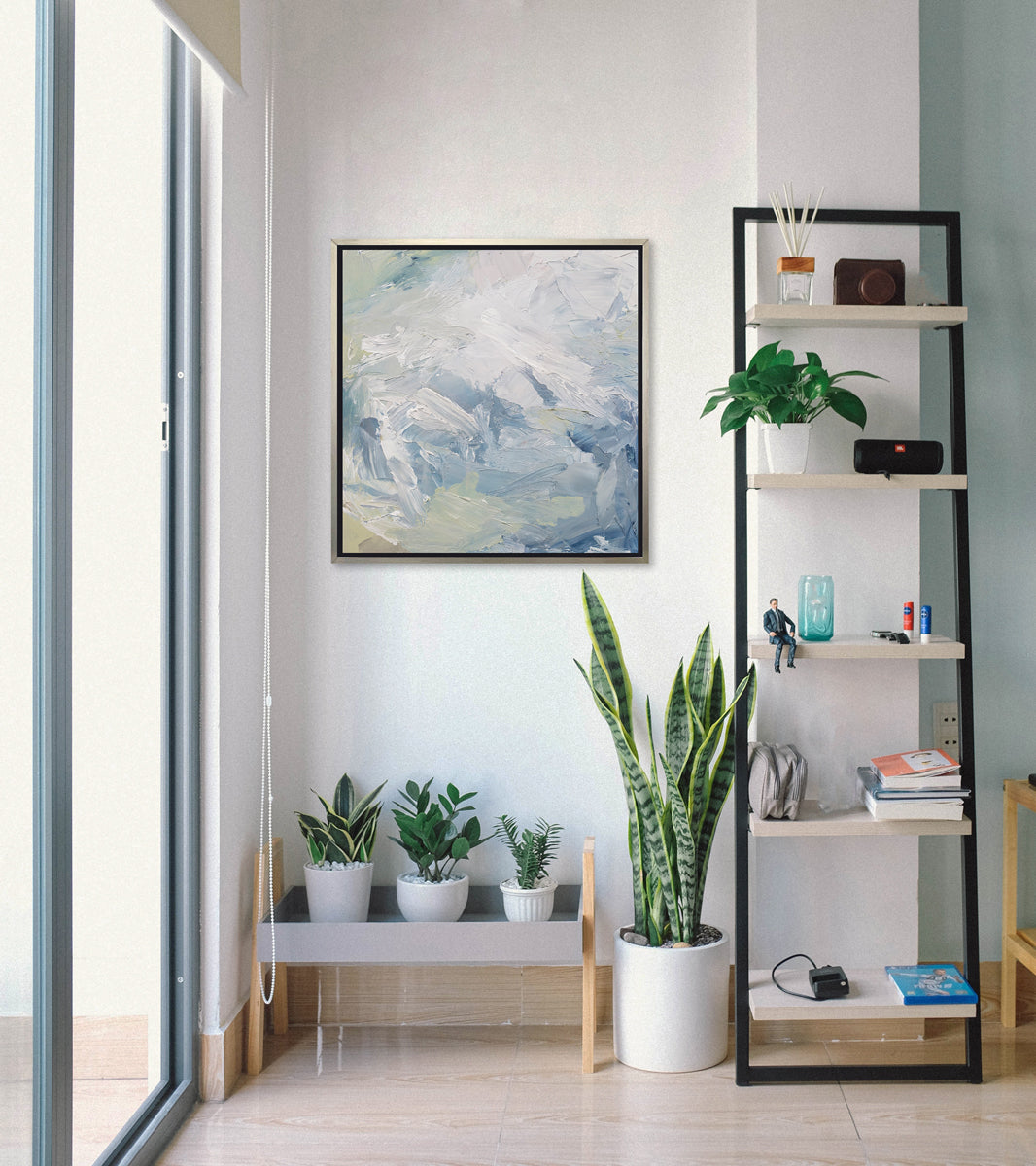 A white, blue, grey and light green abstract print in a silver floater frame hangs on a white wall above a potted plants on a shelf, a larger potted plant and next to a ladder shelf with decorative accessories.