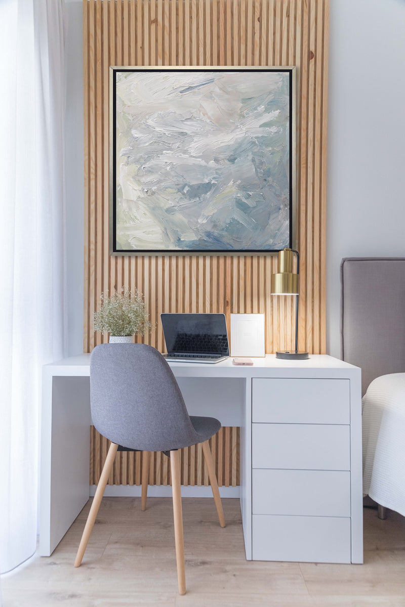 A white, blue, grey and light green abstract print in a silver floater frame hangs on a paneled accent wall above a white desk and grey chair with a laptop computer, a desk lamp and a small potted floral decoration.