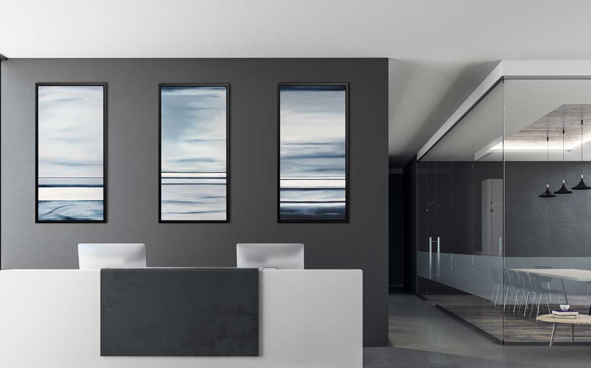 Three blue and white abstract prints hang in a black floater frame on a charcoal grey wall above a white receptionist desk with two desktop computers and a glass encased meeting room to the right.