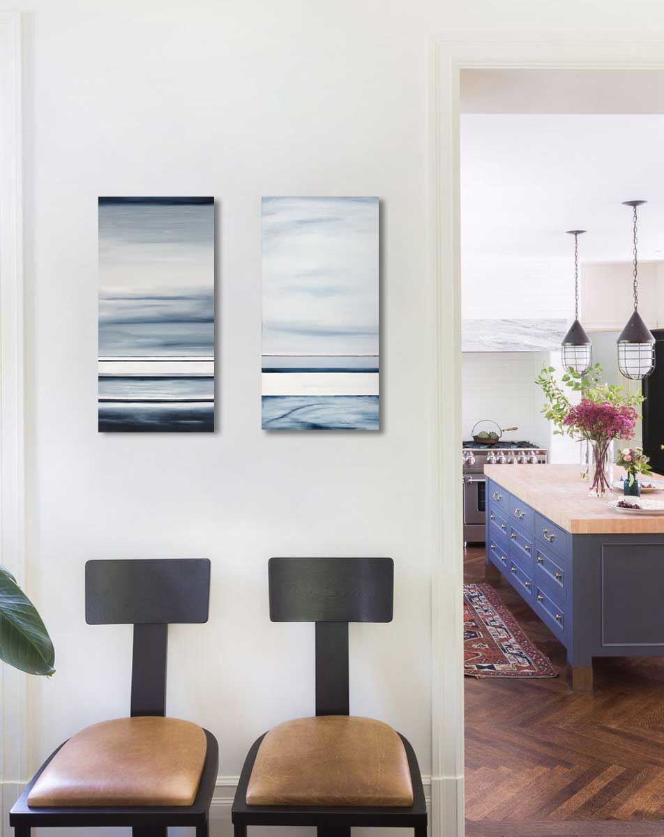 Two vertical paintings of different sized bands of hues of blue and white hang above two dining chairs on a small wall. Next to the wall is an opening into a large kitchen.