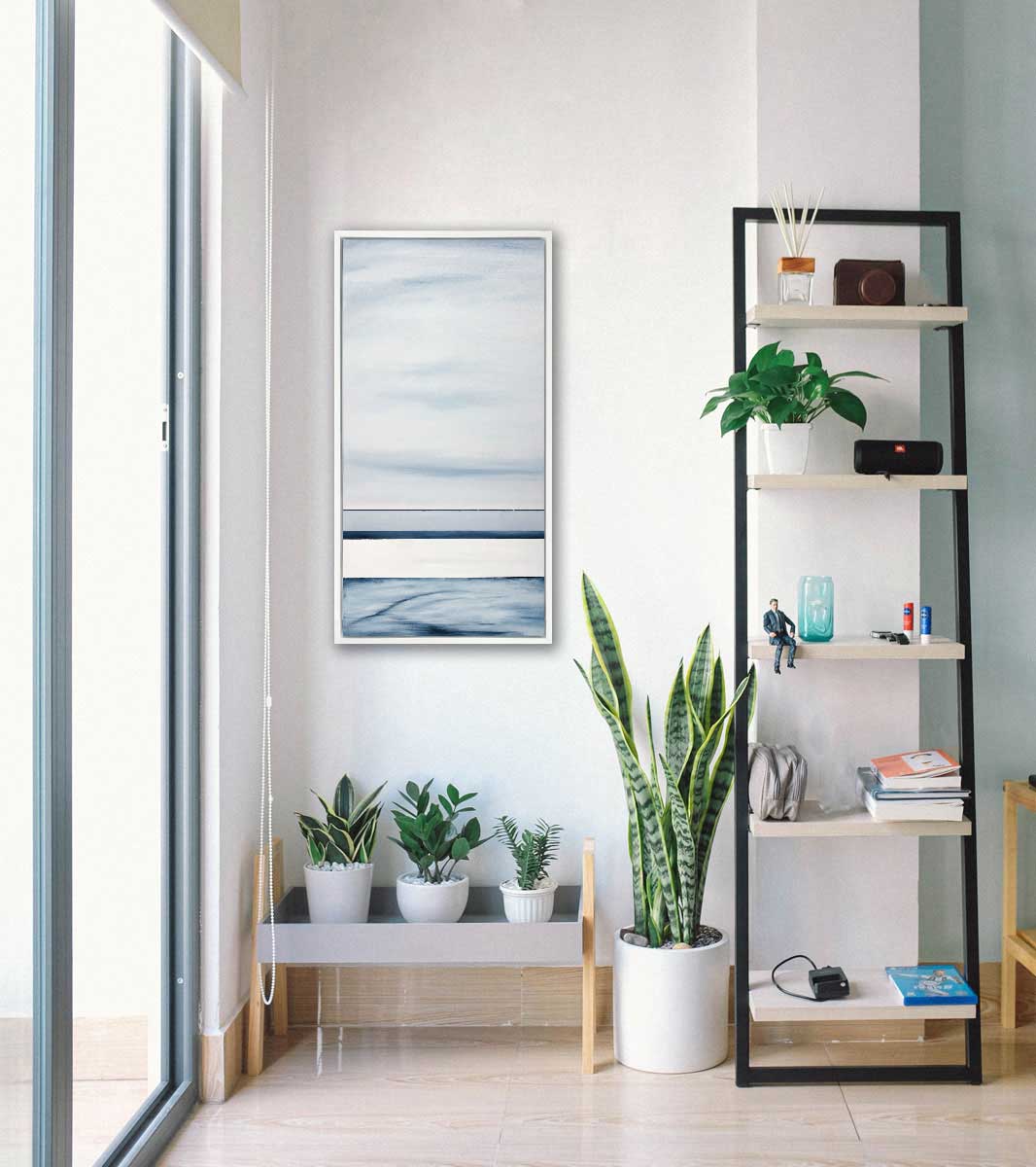 A blue-grey abstract print hangs in a white floater frame above a low shelf with three potted plants next to a larger floor plant and a ladder shelf.