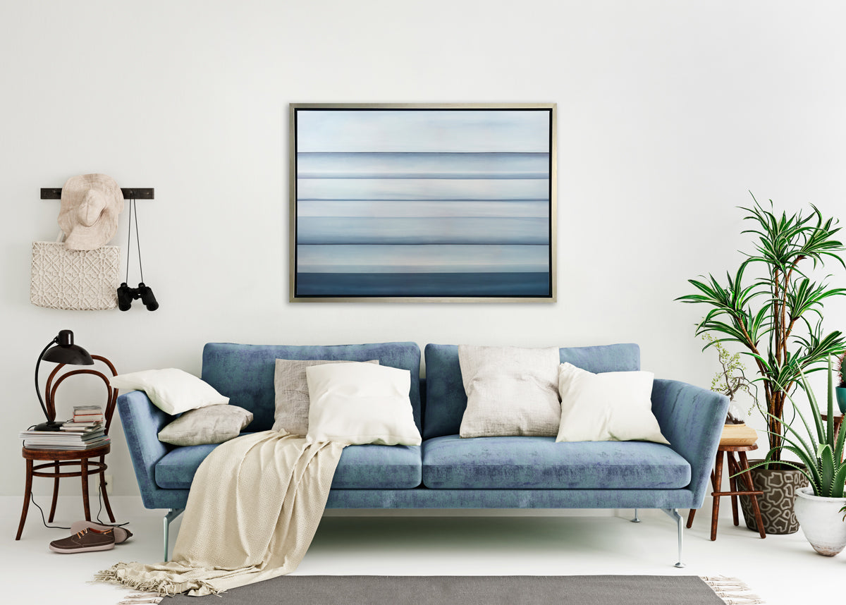 A tonal blue abstract landscape print in a silver floater frame hangs on a white wall above a blue sofa with white pillows, two potted floor plants and a wood chair with a stack of books and a black adjustable table lamp.