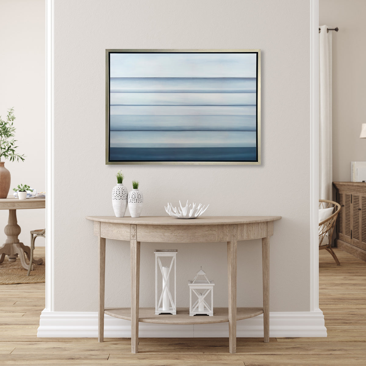 A tonal blue abstract landscape print in a silver floater frame hangs on a beige half wall above a natural wood half circle console table with two white vases, a white decorative bowl and two white lanterns.