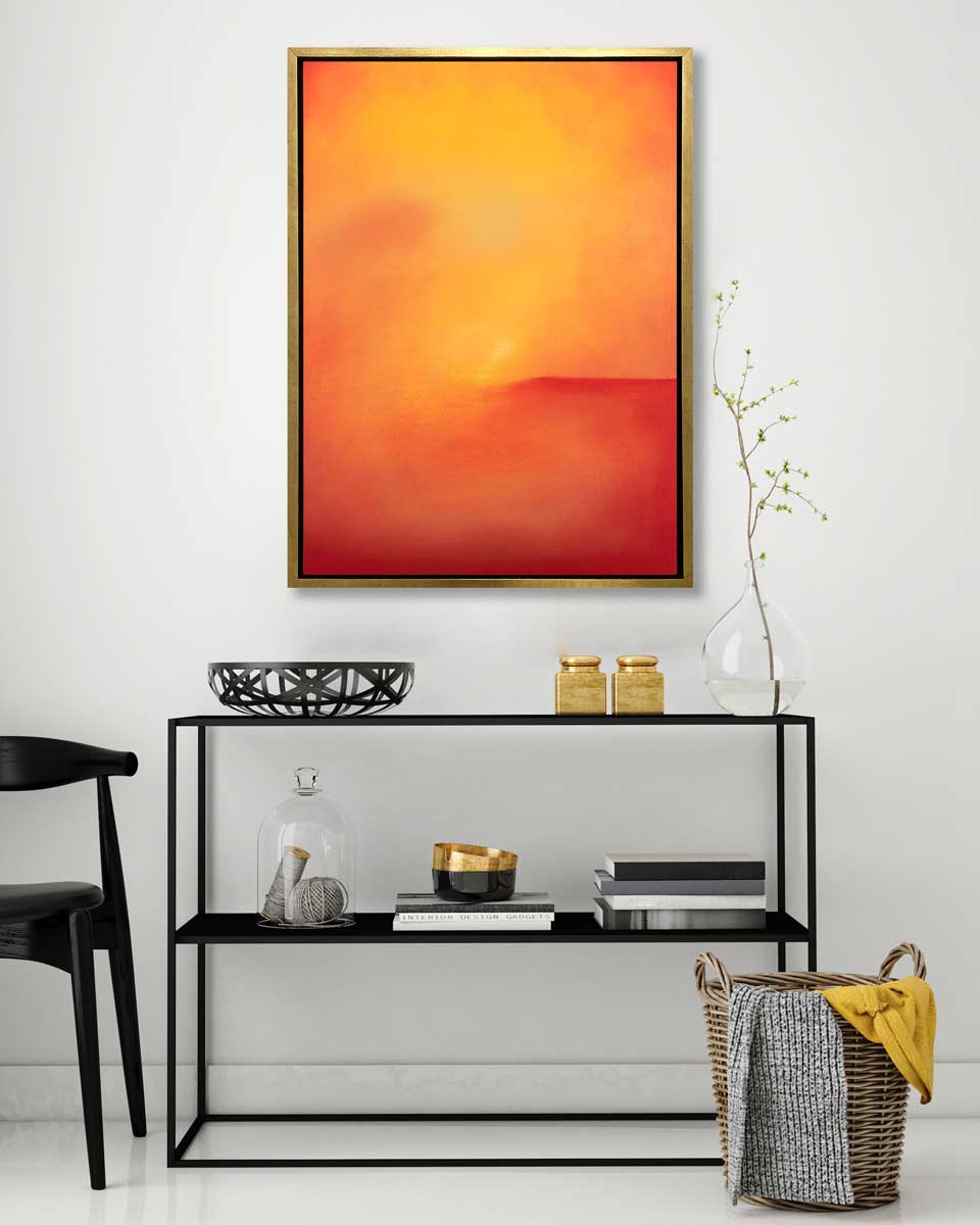An orange and red abstract print in a gold floater frame hangs on a white wall above a black console table with a modern black bowl, stacks of books, two gold containers and a glass vase with a greenery stem.