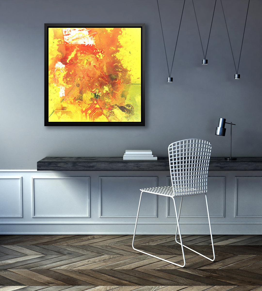 A yellow, orange and white abstract print in a black floater frame hangs on a grey wall above a black floating desk with a stack of books, a small chrome desk lamp and a white chair.