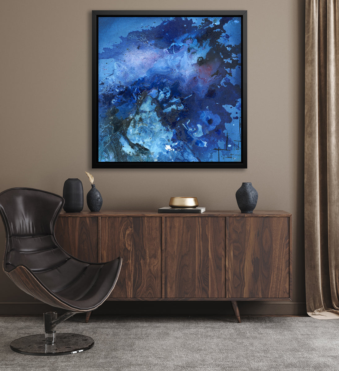 A blue and black abstract print in a black floater frame hangs on a beige wall above a dark wood sideboard with black vases, a gold decorative bowl and a brown leather modern chair.