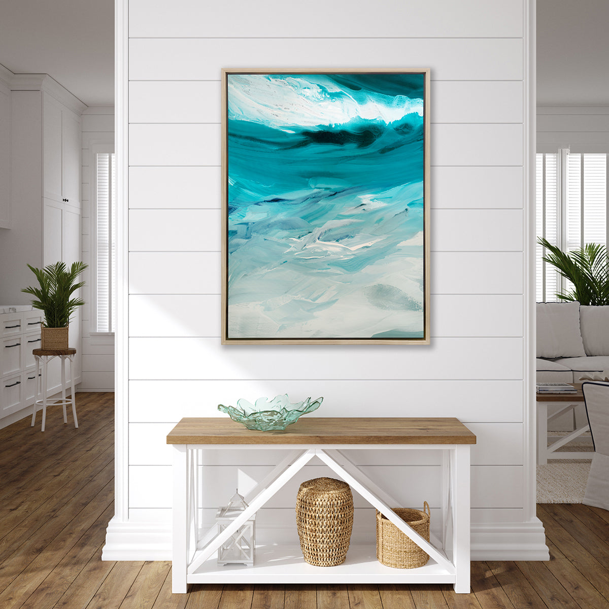A teal, white and grey abstract print in a white oak floater frame hangs on a white wall above a wood console table with a teal glass decorative bowl, a white lantern and two woven baskets.