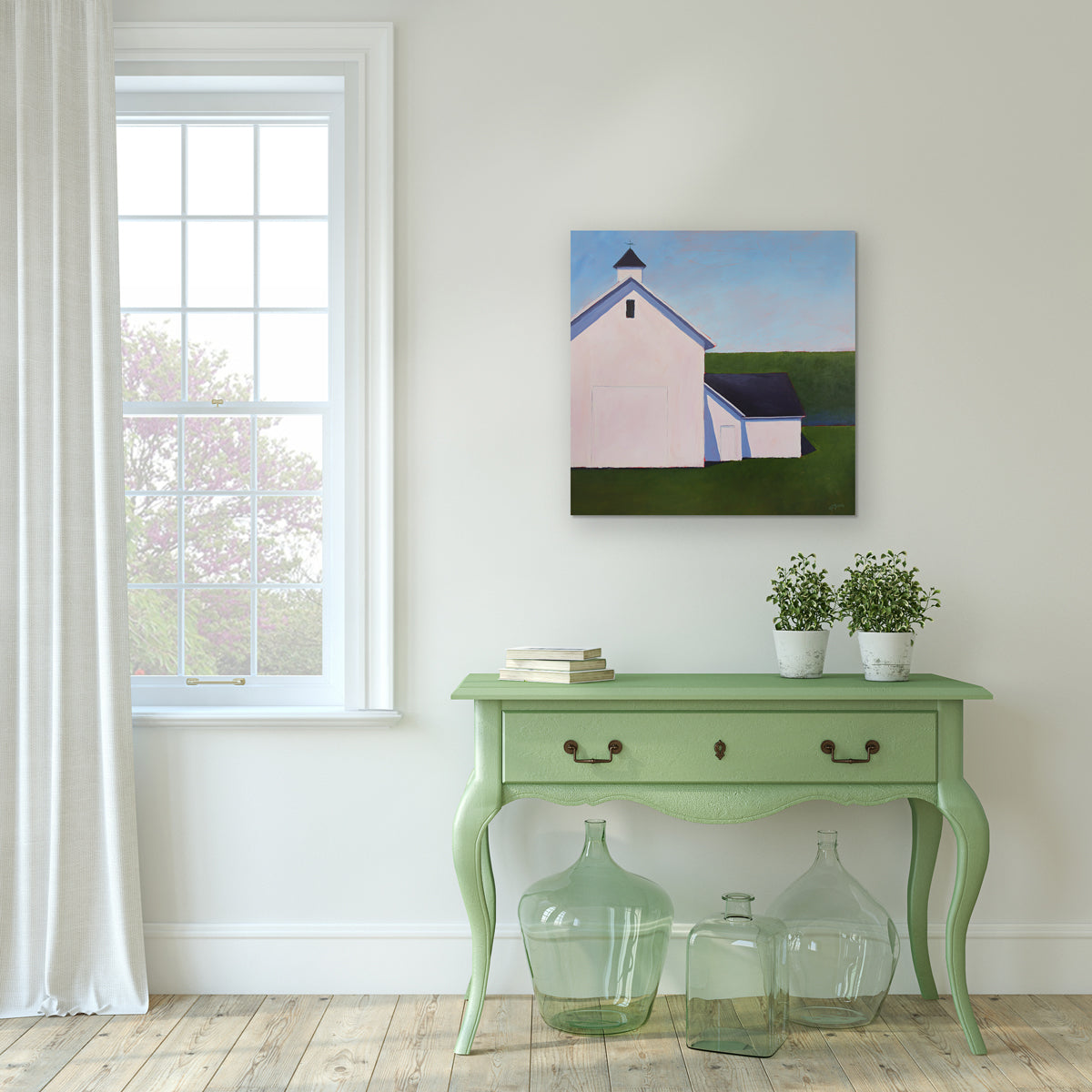 A painting of a white barn with a weather vane in front of a hedge is hung above a table.
