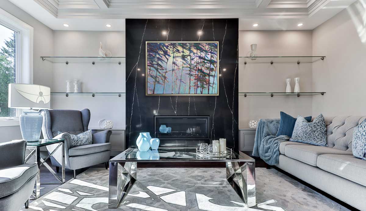 A light blue, green and dark green abstract tree landscape print in a silver floater frame hangs on a black marble wall of a fireplace across from a dove grey sofa, two matching wingback chairs and a chrome square coffee table.
