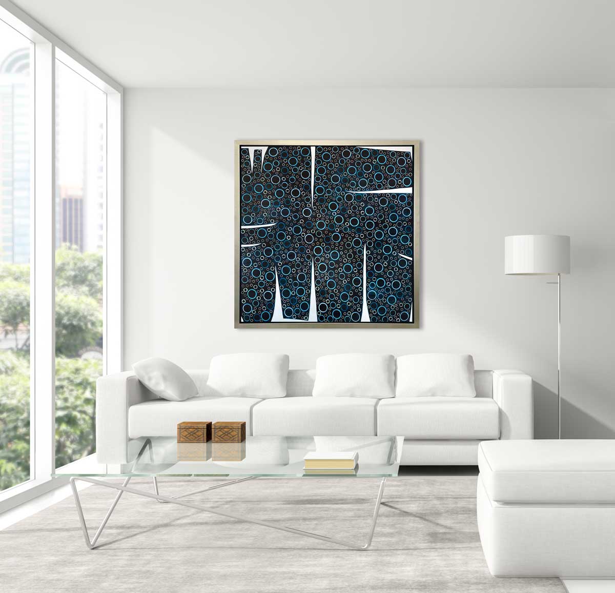 A black, blue and white abstract print in a silver floater frame hangs on a white wall above a white sofa with pillows, a glass coffee table and a floor lamp with a white shade.