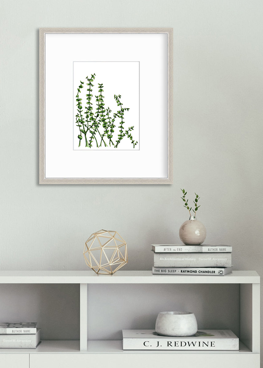 A print of green eucalyptus stems in a silver frame with a mat hangs on a white wall above a white shelf with stacks of books, a gold wire sphere decoration and a small beige ornament vase with greenery stems.