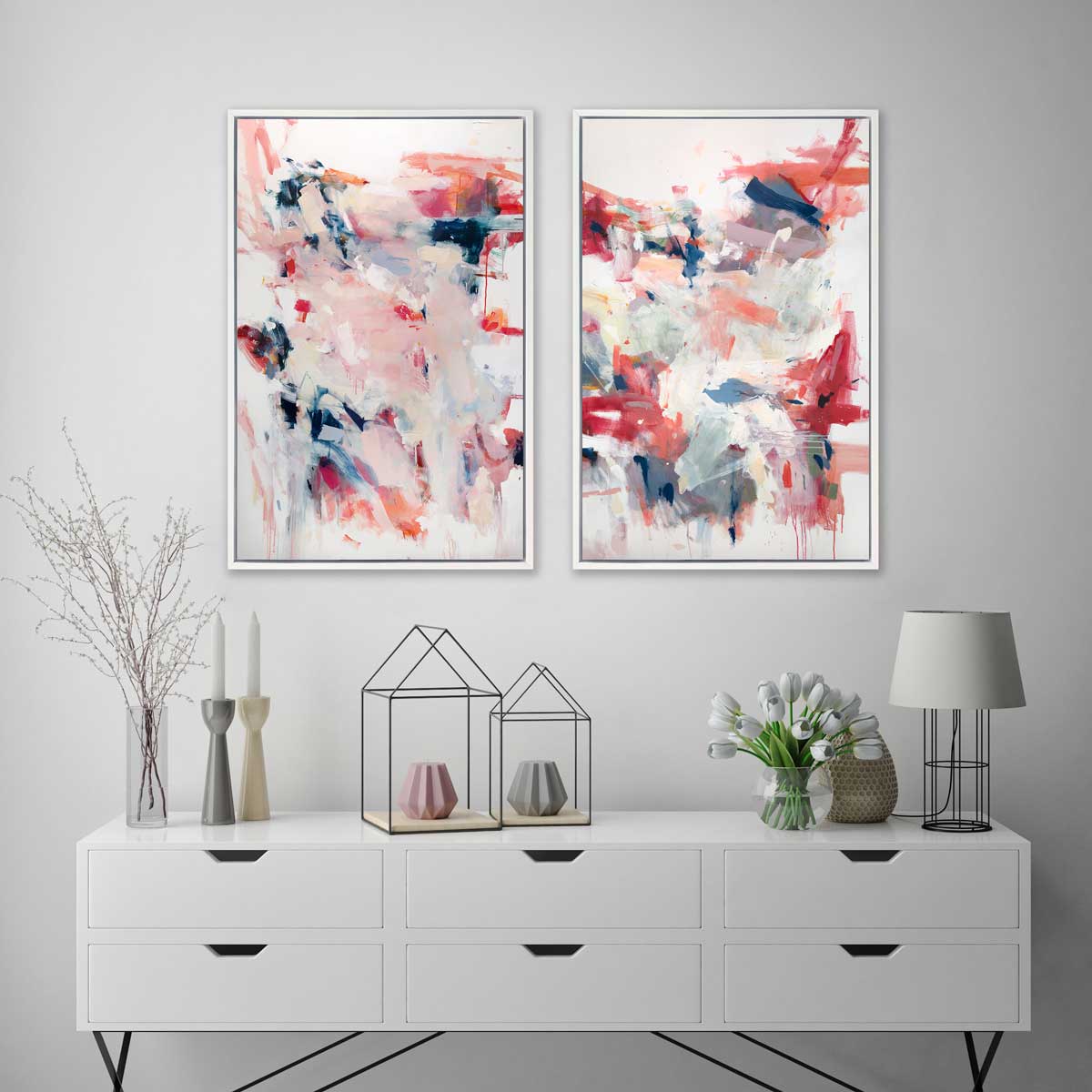 Two pink, blue and white abstract prints in white floater frames hang side by side on a white wall above a white six-drawer desk with candles, a small wire table lamp and a small rounded glass vase with white tulips.