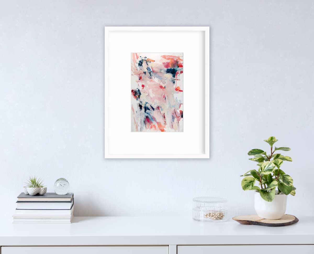 A pink and white abstract print by Kelly Rossetti in a white frame with a mat hangs on a white wall above a white surface with a stack of books, a round clear container with gold accessories and a potted plant.