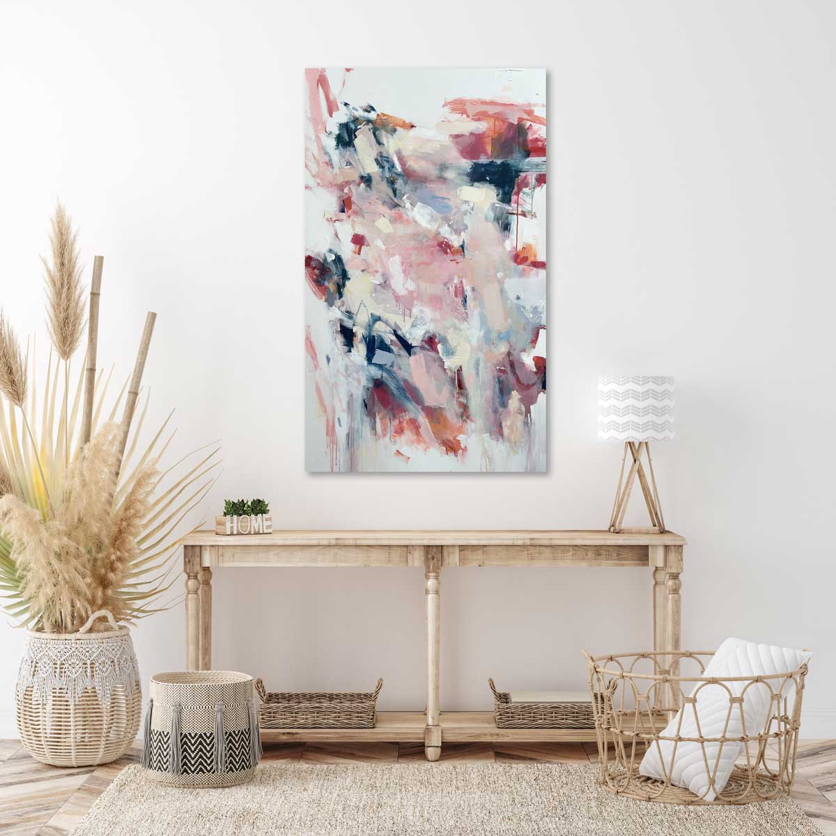 A light pink and white abstract painting by Kelly Rossetti hangs on a white wall above a wooden console table with a table lamp, four woven baskets and a giant woven basket with bamboo stems.