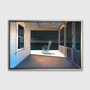 A print of a white rocking chair on a deck of a house with a high contrast light on the chair in a silver floater frame hangs on a white wall.