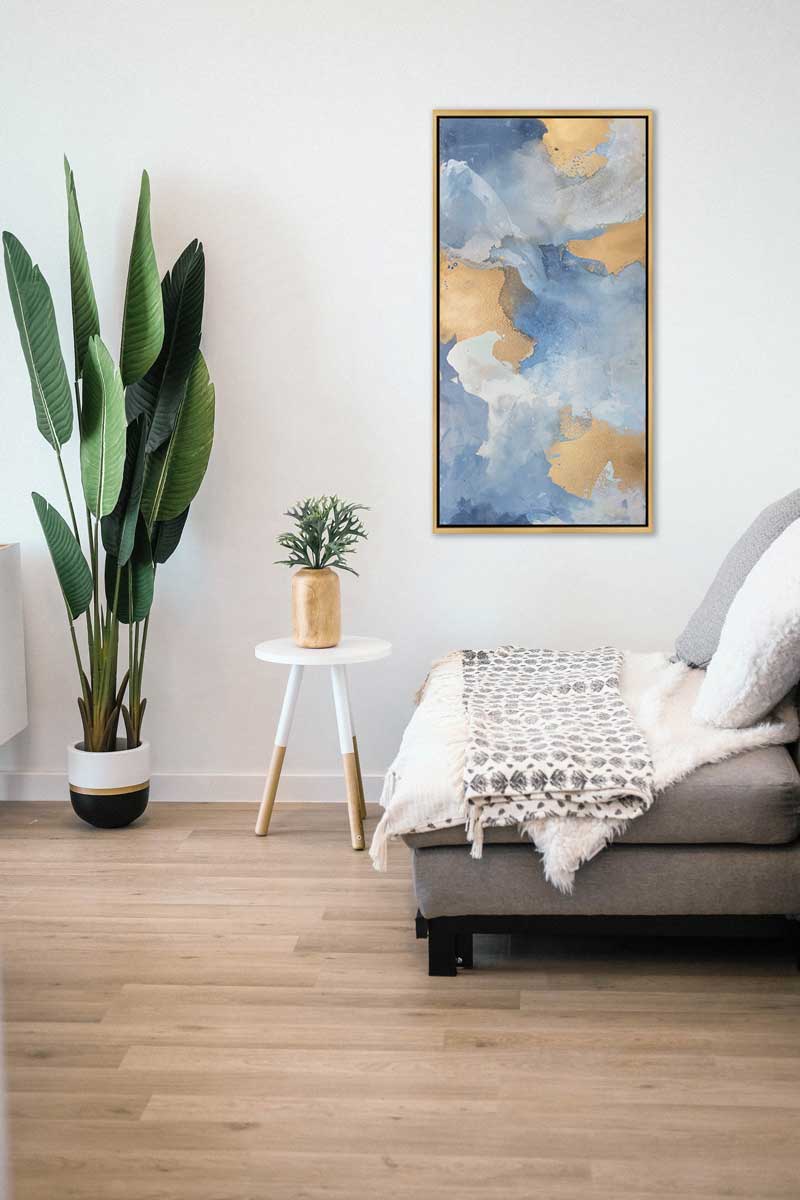A blue, gold and white abstract print in a gold floater frame hangs on a white wall above a small grey sofa with blankets, a white side table with a gold vase and greenery and a tall potted floor plant.