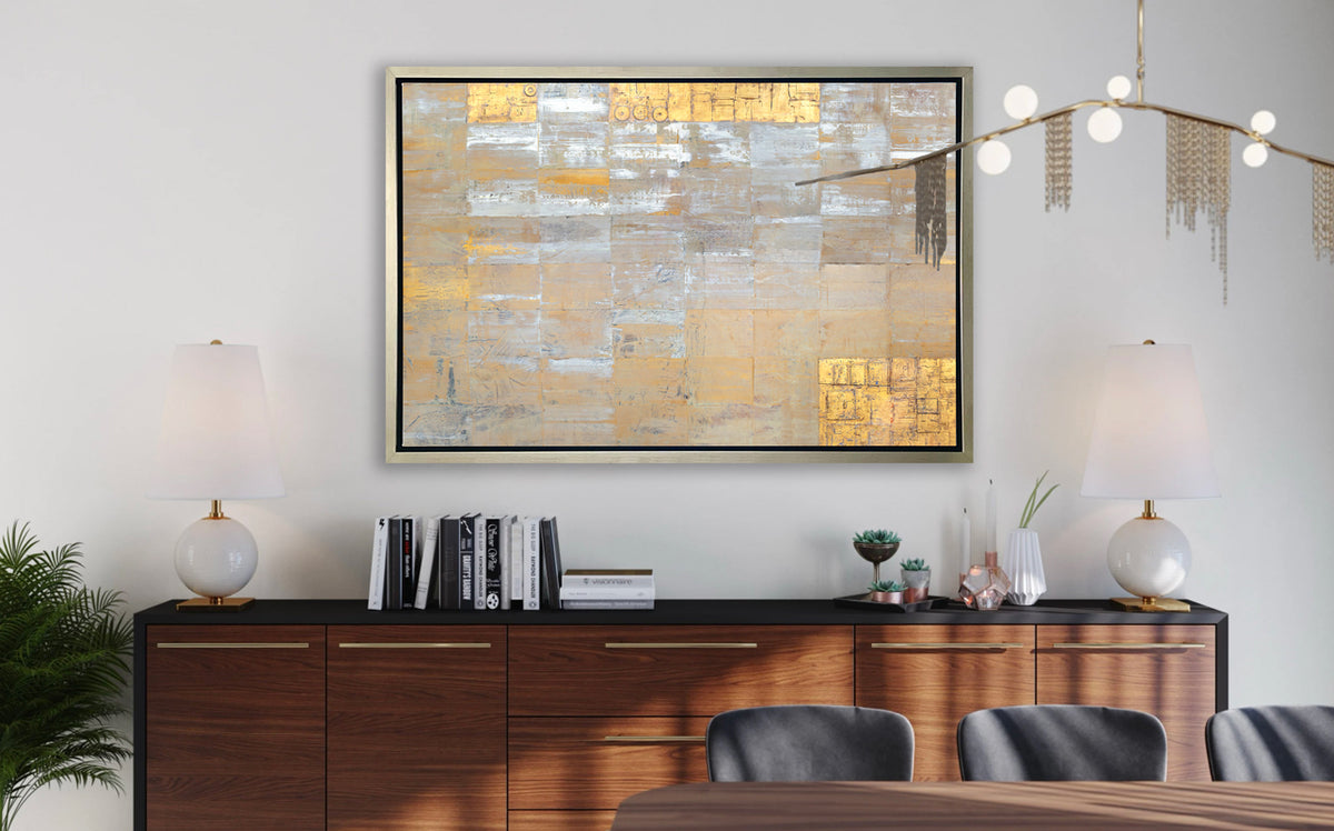 A grey, silver and gold abstract geometric print in a silver floater frame hangs on a white wall above a black and dark wood sideboard with a stack of books, decorative accessories, two white table lamps and a wood table with grey chairs.
