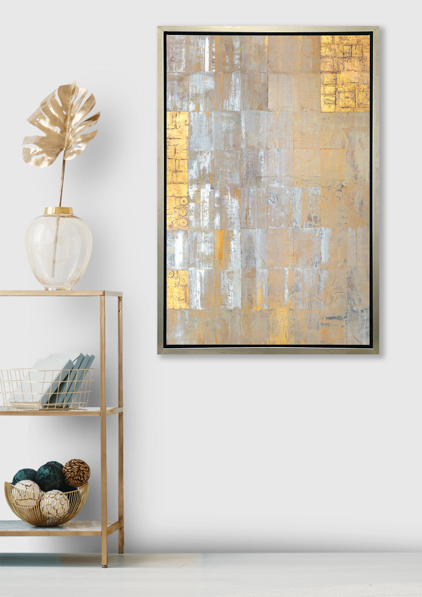 A grey, silver and gold abstract geometric print in a silver floater frame hangs on a white wall next to a gold three-tiered shelf with gold accessories.
