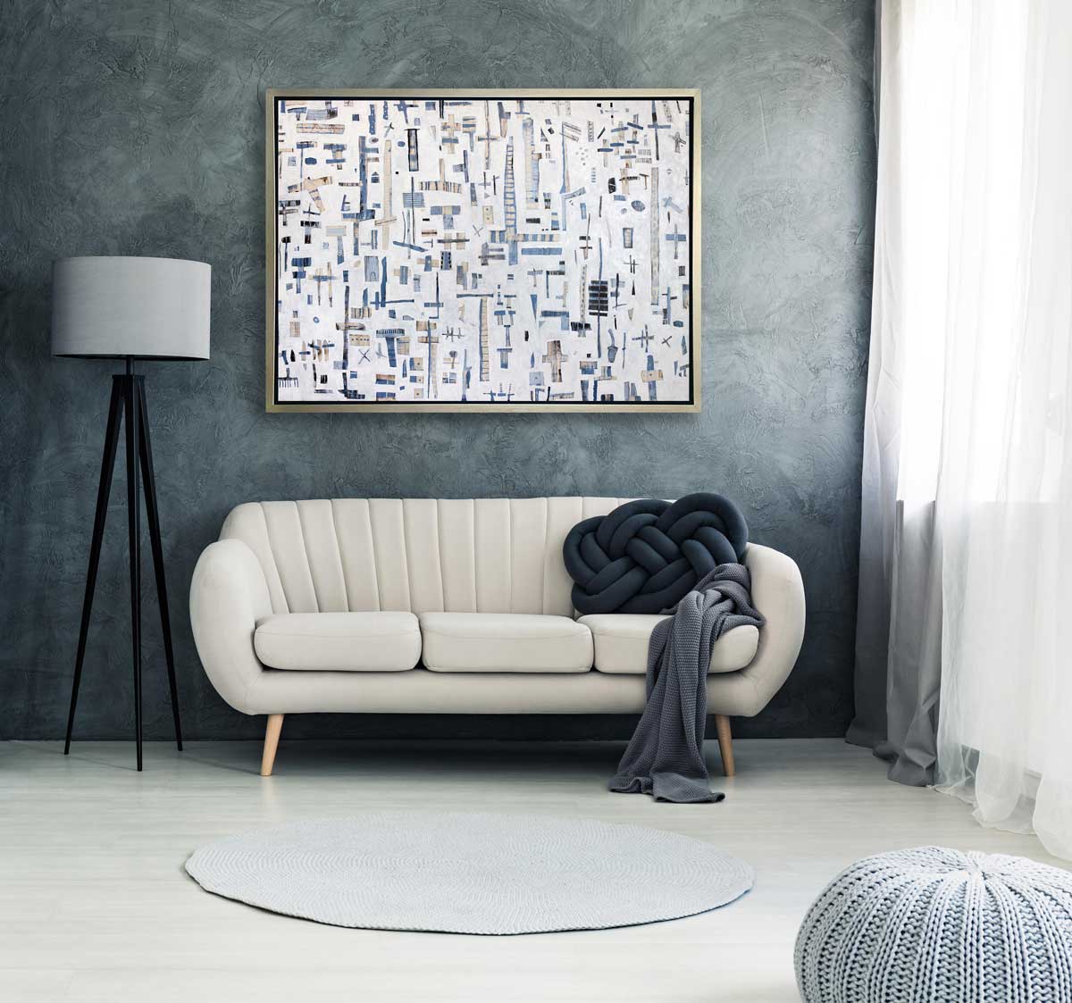 A white, light blue and beige abstract print in a silver floater frame hangs on a grey wall above a white sofa with a woven dark grey pillow, a light grey throw blanket and a black tripod floor lamp.