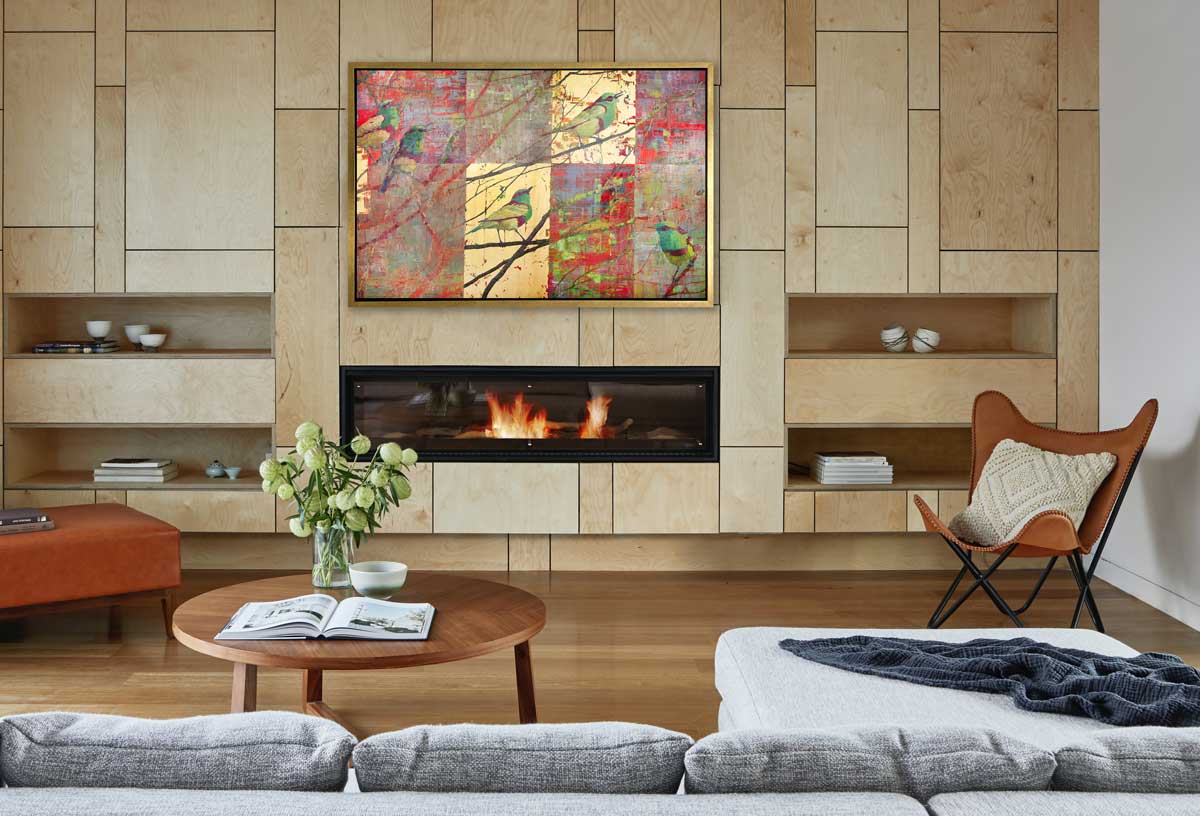 A red, gold and green geometric abstract print of birds in a tree in a gold floater frame hangs on a natural wood panel wall above an wall fireplace insert, built-in shelves, a brown chair and ottoman and a round coffee table.
