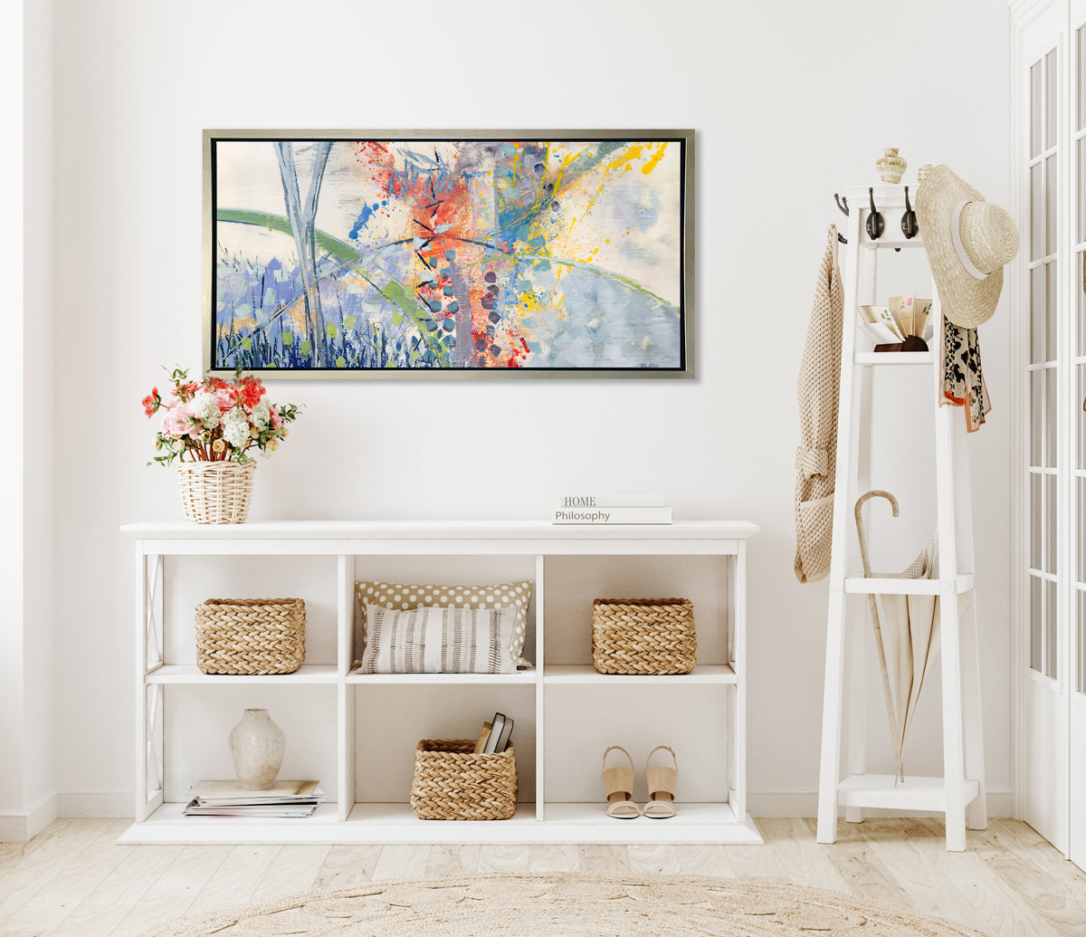 A multicolored abstract print in a silver floater frame hangs on a white wall above a white shelving unit with woven baskets, neutral pillows, a spring floral potted bouquet and a tall white coatrack with accessories.