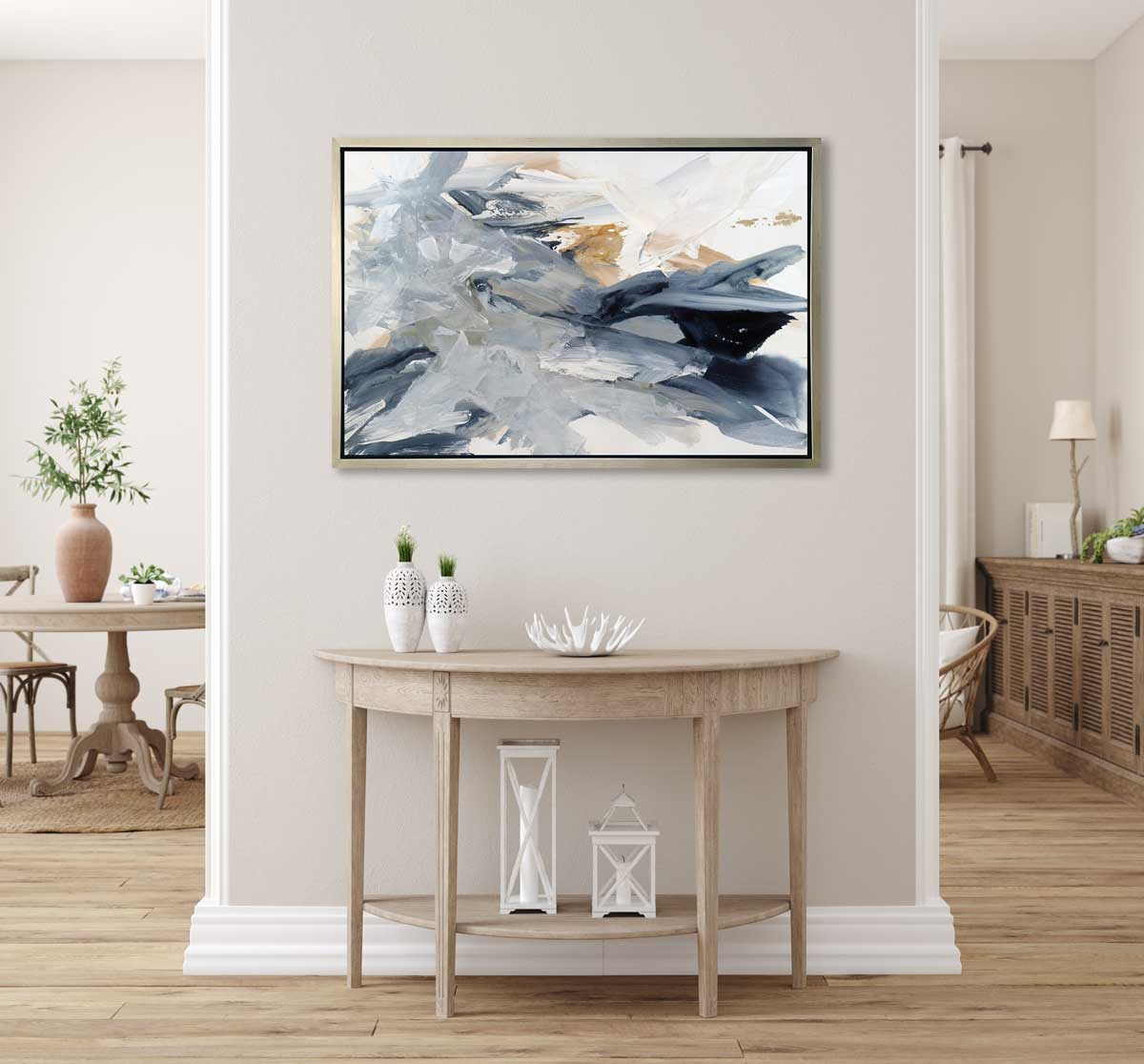 A grey, blue, gold and white abstract print in a silver floater frame hangs on a white wall above a natural wood half-circle console table with two white vases, a white decorative bowl and two white lanterns.