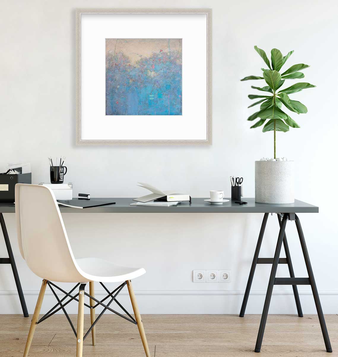 A blue abstract landscape print in a silver frame with a mat hangs on a white wall above a dark grey desk with open notebooks, a black coffee mug with pencils and a potted plant.
