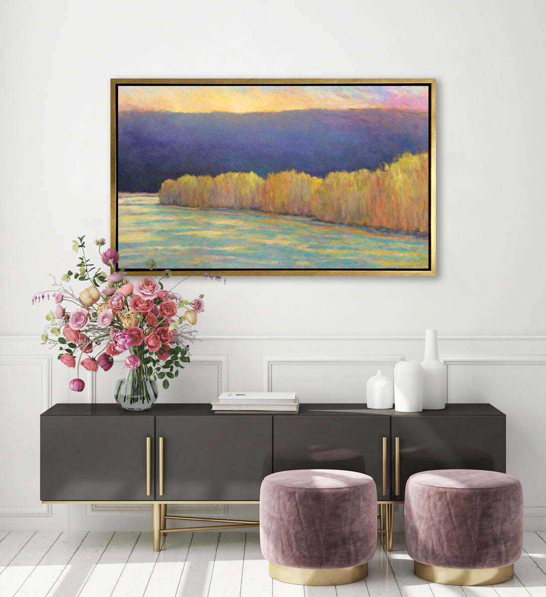 A purple, yellow, orange and blue abstract landscape print in a gold floater frame hangs on a white wall above a black console cabinet with a stack of books, an arrangement of white vases, a glass vase with a colorful flower arrangement and two mauve ottomans on the floor.