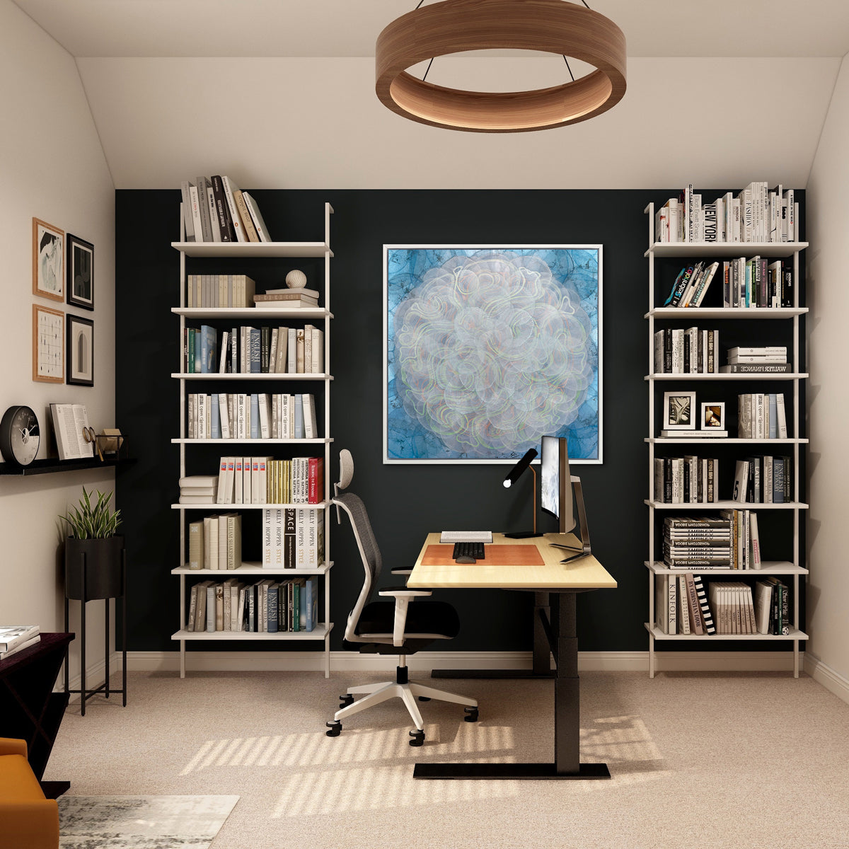 A blue, white, orange and yellow abstract geometric print in a white floater frame hangs on a black wall in between two white bookshelves and above a desk with a computer monitor and desk lamp and a rolling desk chair.